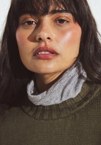 Green knitted sweater with ribbed neckline layered over a gray turtleneck, featuring smooth texture and minimal detailing. Natural lighting highlights skin.