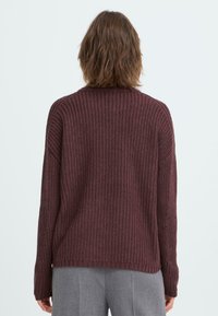 Pull en tricot de couleur bordeaux, présentant un motif en relief, des épaules tombantes et des manches longues avec une coupe décontractée, associé à un pantalon gris.