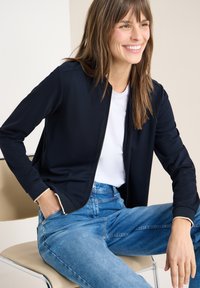 Femme souriante assise sur une chaise beige, portant une veste zippée bleu marine, un t-shirt blanc et un jean taille haute bleu, avec une main dans la poche.
