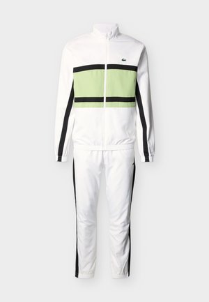 Survêtement blanc avec des bandes noires sur les manches et les jambes, panneau horizontal vert clair sur la veste, et petit logo de crocodile vert sur la poitrine.