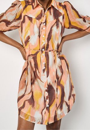 Robe à boutons sur le devant avec un motif abstrait rose, marron, blanc et jaune, manches bouffantes, col et ceinture à nouer à la taille, portée par une personne les mains sur les hanches.