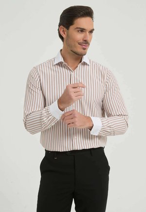 Homme ajustant ses poignets de chemise blanche à rayures brunes et col blanc, portant un pantalon noir, debout devant un fond clair uni.