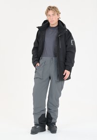 Zwarte waterdichte jack met rits aan de voorkant en een logo op de mouw, gecombineerd met grijze cargo-stijl broek met zijzakken en verstevigde knieën.