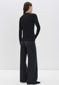 Frau mit langen braunen Haaren, die ein schwarzes langärmliges Oberteil und weite schwarze Jeans trägt, steht mit dem Rücken zu einer schlichten hellen Hintergrund.