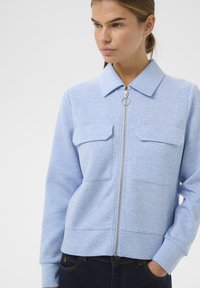 Hellblaues Zip-Up-Jackett aus weichem Stoff, mit einem Kragen, zwei Brusttaschen und gerippten Bündchen sowie Saum.