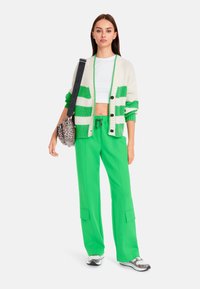 Strickjacke in creme- und grünen Streifen, enges weißes Cropped-Top, grüne weite Hose und Sneakers mit einem Leopardenmuster.