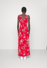 Osoba oblečená v červených květinových maxi šatech s křížovými ramínky na zádech a bílými teniskami, stojící na jednobarevném světlém pozadí.