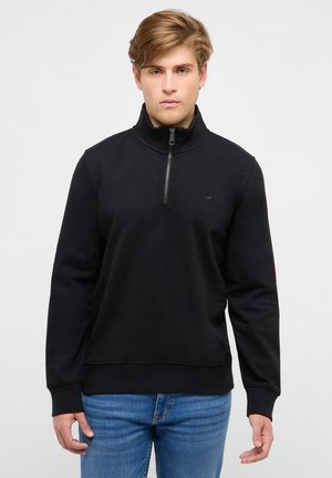 Junger Mann trägt einen schwarzen Pullover mit Reißverschluss bis zur Brust und blaue Jeans, steht vor einem schlichten hellen Hintergrund.