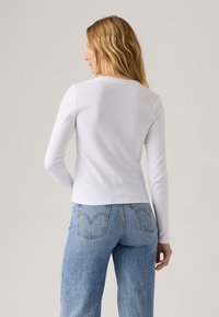 Bílé tričko s dlouhým rukávem a kulatým výstřihem, přiléhavý střih. Kombinováno se světle modrými džínami s výrazným šitím na zadních kapsách.