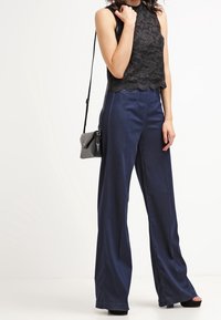 Top con pizzo nero a halter con orlo smerlato abbinato a pantaloni larghi blu navy. Il modello tiene una piccola borsa a tracolla nera.