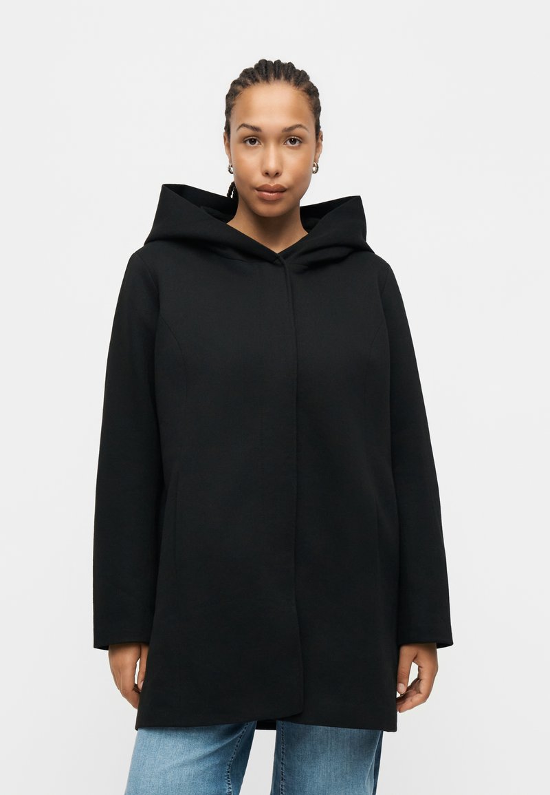 Woman stands facing forward, wearing a black hooded coat and blue jeans against a plain light background. Hands relaxed at sides.