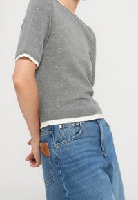 Suéter gris de manga corta con ribete blanco y pequeños adornos dispersos, combinado con jeans de mezclilla azul de cintura alta que presentan un parche de cuero.