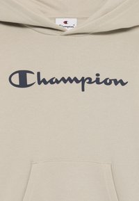 Vaaleanruskea huppari pehmeästä kankaasta, jossa on tumma merimetsovärinen "Champion" -logo rinnassa, etupocket ja nauhakuori.