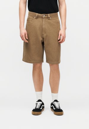 Homme portant un short marron arrivant aux genoux, des chaussettes blanches, des baskets noires et blanches, et un t-shirt noir, debout devant un fond uni.