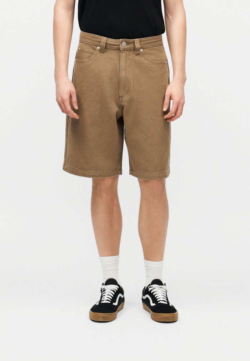 Άνδρας που φοράει καφέ βερμούδα μέχρι το γόνατο, λευκές κάλτσες, μαύρα και άσπρα αθλητικά παπούτσια και μαύρο πουκάμισο, στέκεται μπροστά σε απλό φόντο.