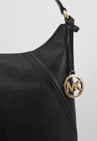 Coin d'un sac à main en cuir noir texturé avec une fermeture éclair dorée et un pendentif rond à logo MK suspendu accroché à la boucle de la sangle.