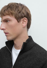 Sweat à fermeture éclair gris foncé avec un col montant, texture côtelée et débardeur blanc contrastant. Profil latéral du modèle sur un fond neutre.