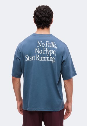 Uomo con capelli corti e ricci che indossa una maglietta blu con la scritta "No Frills, No Hype, Start Running" sulla schiena, su sfondo semplice.