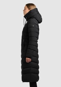 Lungo cappotto imbottito nero con cappuccio, dotato di un design a righe, tasche laterali e un logo sulla manica sinistra. Tessuto liscio e isolato.