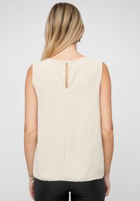 Bluză bej fără mâneci cu o suprafață texturată, având închidere la spate tip clepsidră și un guler rotund. Design simplu, minimalist.