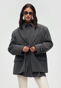 Donna in un blazer grigio scuro oversize e gonna, indossando grandi orecchini d'argento, occhiali da sole scuri, anello d'argento e pantaloni avorio.