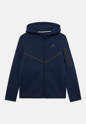 Sweat-shirt à capuche bleu marine avec une fermeture éclair intégrale, featuring une bande noire diagonale sur la poitrine et une poche latérale zippée. Logo Nike sur la poitrine.
