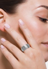 Sølv ribbet ring med et glatt, buet design. Båret på en finger med lyserosa polerte negler, mot en nøytral hudtonebakgrunn.