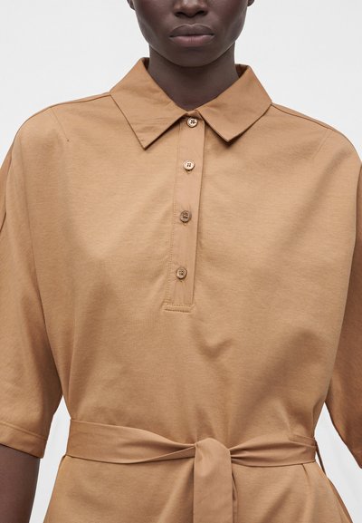 Personne à la peau foncée portant une robe marron clair à col avec manches courtes, patte de boutonnage et ceinture en tissu assorti nouée à la taille.