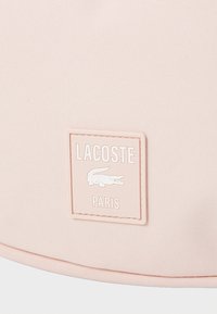 Artimas vaizdas, kuriame matyti šviesiai rožinė tekstūruota medžiaga su siūtu kvadrato etiku, kuriame parašyta "LACOSTE PARIS", ir įspaustu krokodilo logotipu.