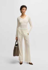 Cream ribbed long-sleeve top with a v-neck paired with cream wide-leg trousers and brown heeled pumps, holding a brown tote bag.