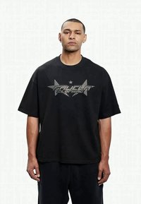 Svart oversized t-skjorte laget av bomull, med sølvstjerne-grafikk og tekstdetalj. Korte ermer, rund hals, avslappet passform.