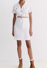 Robe blanche à manches courtes avec un devant à boutons, deux poches poitrine et une ceinture marron. Présente une coupe droite et un tissu texturé.