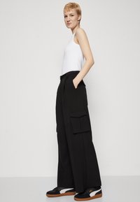 Black wide-leg cargo pants with two large side pockets, high waist, and a sleek finish, paired with a white tank top and black sneakers.