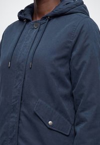 Personne portant une veste à capuche bleu marine avec cordons, une fermeture à boutons près du cou et une poche avant avec rabat à pression.