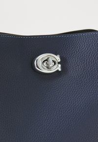 Gros plan sur un fermoir à tour argenté sur un sac en cuir bleu foncé texturé avec une couture bleue visible le long du bord supérieur.