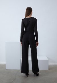 Un haut noir à manches longues associé à un pantalon large noir bien ajusté et des talons hauts, le tout sur un fond minimaliste avec des formes géométriques.