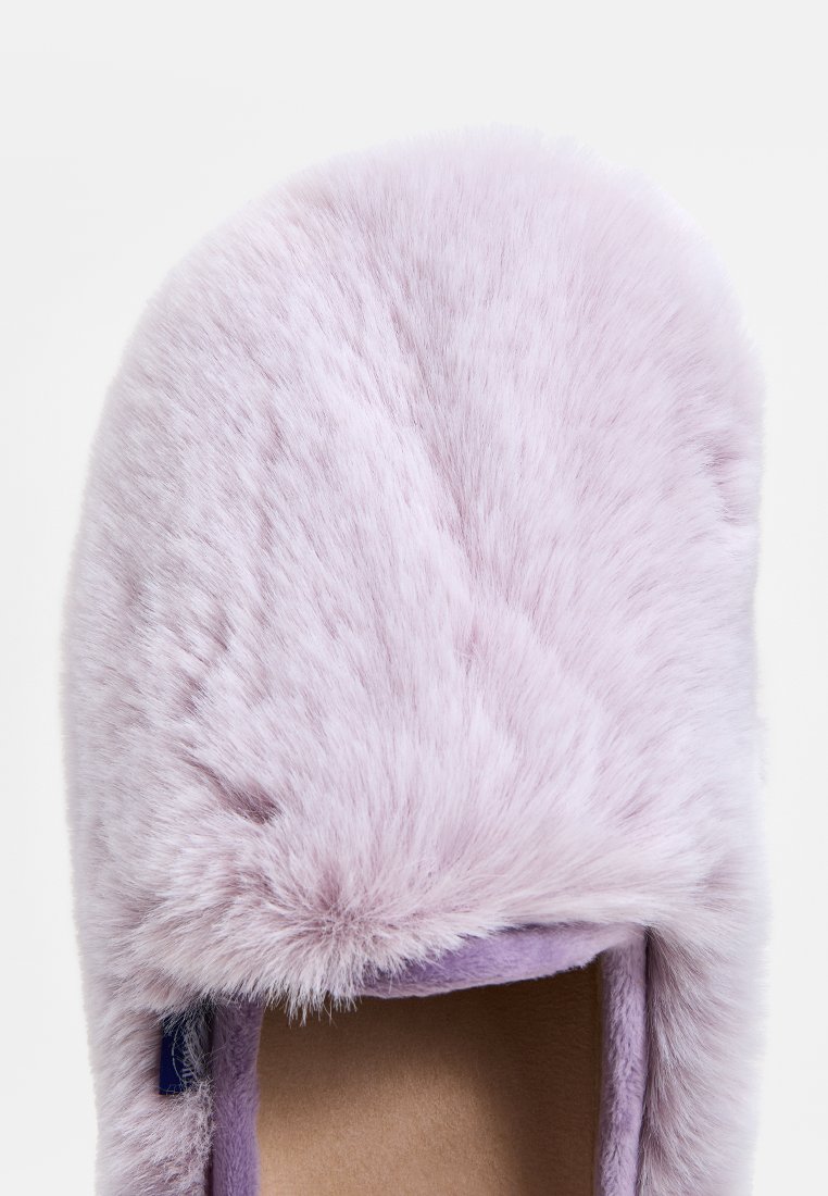 Lavendelfarbenes Slipper mit weichem, langem Kunstfellmaterial, abgerundeter Zehenform und einer strukturierten Oberfläche mit einem kleinen, sichtbaren Markenetikett.