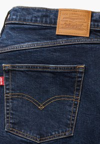 Jeans en denim foncé avec une poche arrière présentant un motif de couture distinct, et une patch en cuir tan avec le logo de la marque et des détails. Étiquette rouge visible.