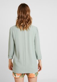 Mintunvihreä kevyt pusero, jossa on kolmen neljänneksen hihat, hienovarainen tekstuuri ja suora leikkaus, puettu kuvioitujen oranssien shortsien päälle.