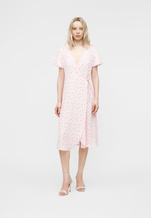 Kvinne står iført en lys rosa omslagskjole med korte flagrende ermer og liten, delikat blomsterprint, kombinert med beige høye hæler.