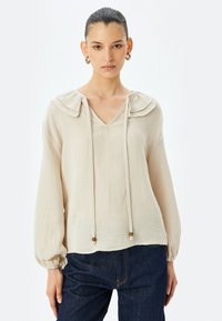 Beige blouse met gerimpelde kraag, V-halslijn, lange poffende mouwen, verstelbare trekkoord met houten kralen, en een structuurstofafwerking.