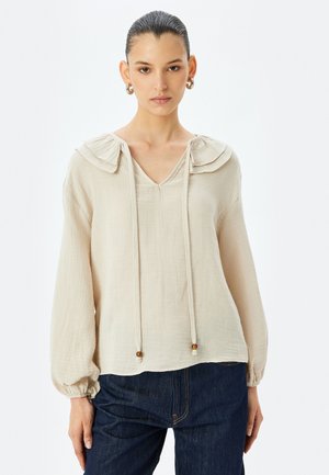 Beige blouse met gerimpelde kraag, V-halslijn, lange poffende mouwen, verstelbare trekkoord met houten kralen, en een structuurstofafwerking.