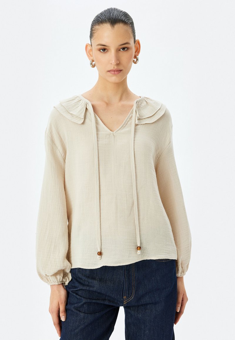 Beige blouse met gerimpelde kraag, V-halslijn, lange poffende mouwen, verstelbare trekkoord met houten kralen, en een structuurstofafwerking.
