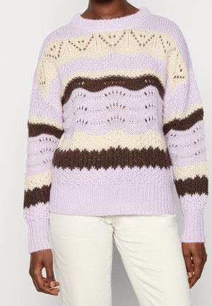 Personne portant un pull en tricot rayé lilas, crème et marron avec un motif texturé ajouré, et un pantalon blanc.