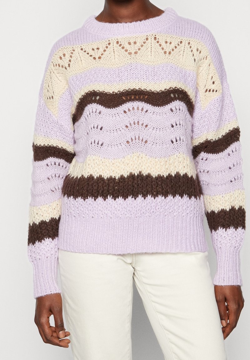 Personne portant un pull en tricot rayé lilas, crème et marron avec un motif texturé ajouré, et un pantalon blanc.