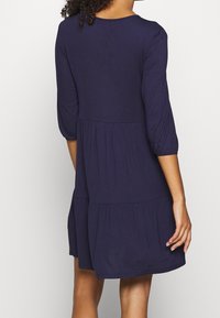 Kvinna i en knälång mörkblå klänning med 3/4 ärmar, som står med ryggen mot kameran framför en helt vit bakgrund.