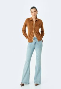 Camisa marrón de botones con cuello, combinada con unos jeans acampanados de color azul claro. Los accesorios incluyen pendientes de aro y zapatos de estampado de leopardo.