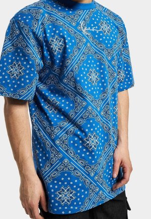 Blaues kurzärmeliges Hemd mit weißen und hellblauen Paisley- und Blumen-Bandana-Mustern, getragen von einer Person, die mit entspannten Händen steht.