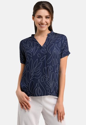 Navy blouse with a v-neckline, short sleeves, and white abstract leaf patterns. Soft fabric with a relaxed fit. Paired with light textured trousers.