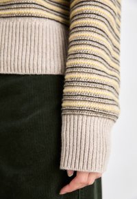Gestreifter Pullover aus beigem, grauem und gelbem Garn. Rippstrickbündchen; strukturierter Strick. Kombiniert mit dunkelgrünem Cordstoff im unteren Bereich.